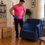 A Summer Moving crew member inspects a blanket-wrapped armchair and labeled boxes in a New York City apartment before pickup.