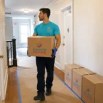 A Summer Moving crew member carries a branded box down an apartment hallway past a stack of packed boxes.